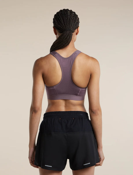 Rear view of a woman wearing a mauve-purple athletic sports bra and black athletic shorts against a plain beige studio background. - Icebreaker Merino Blend 125 ZoneKnit Racerback Bra (Women's) - Grey Plum