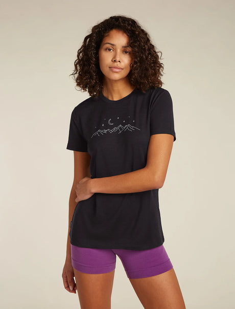 A woman modeling a black short-sleeve t-shirt with a white mountain and moon graphic on the chest, paired with purple athletic shorts, against a solid light beige background. - Icebreaker Merino 150 Tech Lite Short Sleeve T-Shirt Sparkling Stars (Women's) - Black