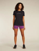 A woman stands against a plain beige studio background wearing a black short-sleeve athletic T-shirt with a small white mountain-and-moon graphic on the chest, purple fitted shorts, and black running shoes. - Icebreaker Merino 150 Tech Lite Short Sleeve T-Shirt Sparkling Stars (Women's) - Black