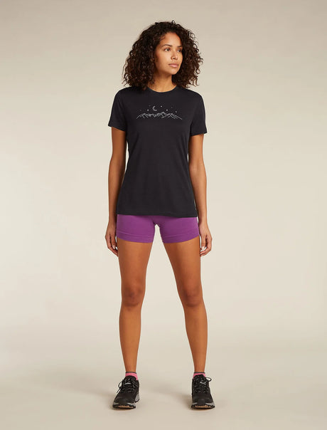 A woman stands against a plain beige studio background wearing a black short-sleeve athletic T-shirt with a small white mountain-and-moon graphic on the chest, purple fitted shorts, and black running shoes. - Icebreaker Merino 150 Tech Lite Short Sleeve T-Shirt Sparkling Stars (Women's) - Black