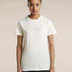 A woman modeling a short-sleeve crewneck t-shirt in off-white, featuring a subtle light brown line-art graphic of mountains and stars across the chest. She is wearing dark pants against a solid, warm beige studio background. - Icebreaker Merino 150 Tech Lite Short Sleeve T-Shirt Sparkling Stars (Women's) - Ecru Heather