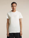 A woman modeling a short-sleeve crewneck t-shirt in off-white, featuring a subtle light brown line-art graphic of mountains and stars across the chest. She is wearing dark pants against a solid, warm beige studio background. - Icebreaker Merino 150 Tech Lite Short Sleeve T-Shirt Sparkling Stars (Women's) - Ecru Heather