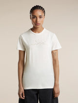 A woman modeling a short-sleeve crewneck t-shirt in off-white, featuring a subtle light brown line-art graphic of mountains and stars across the chest. She is wearing dark pants against a solid, warm beige studio background. - Icebreaker Merino 150 Tech Lite Short Sleeve T-Shirt Sparkling Stars (Women's) - Ecru Heather