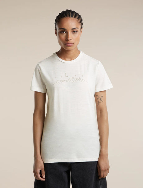 A woman modeling a short-sleeve crewneck t-shirt in off-white, featuring a subtle light brown line-art graphic of mountains and stars across the chest. She is wearing dark pants against a solid, warm beige studio background. - Icebreaker Merino 150 Tech Lite Short Sleeve T-Shirt Sparkling Stars (Women's) - Ecru Heather