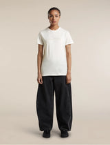 A model stands against a plain beige studio background wearing a white short-sleeve t-shirt with a subtle tonal graphic on the chest, paired with loose black wide-leg jeans and black shoes. - Icebreaker Merino 150 Tech Lite Short Sleeve T-Shirt Sparkling Stars (Women's) - Ecru Heather