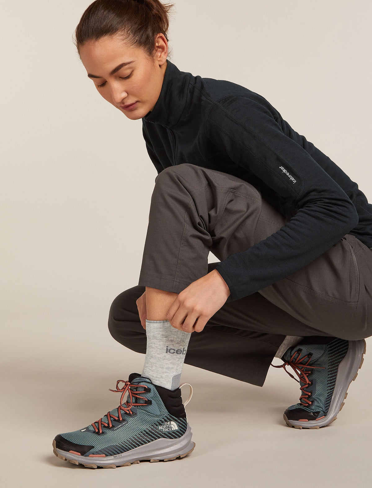 A woman crouches while putting on a gray sock, wearing a black fleece jacket, dark gray pants, and dark teal trail running shoes with orange laces against a light beige studio background. - Icebreaker Merino Hike+ Light Crew Socks (Women's) - Blizzard Heather/Alloy