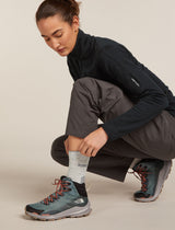 A woman crouches while putting on a gray sock, wearing a black fleece jacket, dark gray pants, and dark teal trail running shoes with orange laces against a light beige studio background. - Icebreaker Merino Hike+ Light Crew Socks (Women's) - Blizzard Heather/Alloy