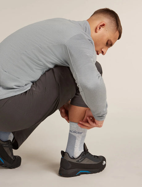 A man crouching down to show off a light gray crew sock with 'baker' printed on it, paired with a light gray long-sleeve shirt, dark gray shorts, and black athletic shoes with blue accents. - Icebreaker Merino Hike+ Medium Crew Socks (Men's) - Blizzard Heather/Alloy
