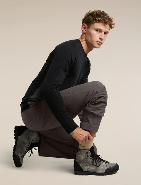 A male model crouching in a studio setting, wearing a black long-sleeve shirt, dark gray pants, and gray hiking boots. He is adjusting his sock, with the brand name 'Kipsta' visible on the cuff. - Icebreaker Merino Hike+ Medium Crew Socks (Men's) - Cumin/Obsidian