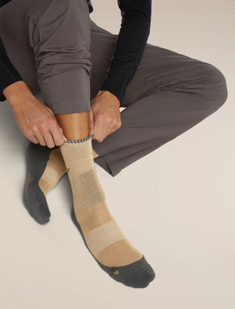 A person sitting and pulling up a striped sock while wearing gray athletic pants and a dark long-sleeve top. The sock has beige and tan horizontal stripes with a dark gray cuff, heel, and toe. - Icebreaker Merino Hike+ Medium Crew Socks (Men's) - Cumin/Obsidian