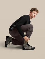 A male model crouching in a studio setting, wearing a black long-sleeve shirt, dark brown pants, and gray high-top sneakers against a solid beige background. - Icebreaker Merino Hike+ Light Crew Socks (Men's) - Cumin/Obsidian
