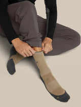 A person sitting and pulling up a tan crew sock with dark gray toes and heels, wearing dark gray pants and a black long-sleeve top, against a plain beige background. - Icebreaker Merino Hike+ Light Crew Socks (Men's) - Cumin/Obsidian
