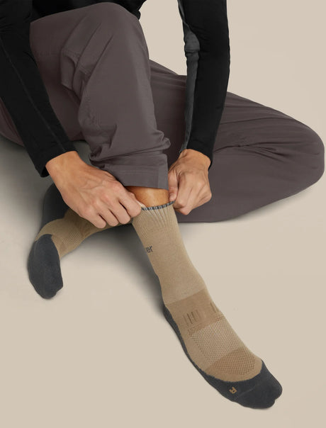A person sitting and pulling up a tan crew sock with dark gray toes and heels, wearing dark gray pants and a black long-sleeve top, against a plain beige background. - Icebreaker Merino Hike+ Light Crew Socks (Men's) - Cumin/Obsidian
