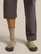 Close-up of a person's lower legs wearing beige crew socks with dark gray toes and heels, paired with dark gray pants, against a solid light beige background. - Icebreaker Merino Hike+ Light Crew Socks (Men's) - Cumin/Obsidian