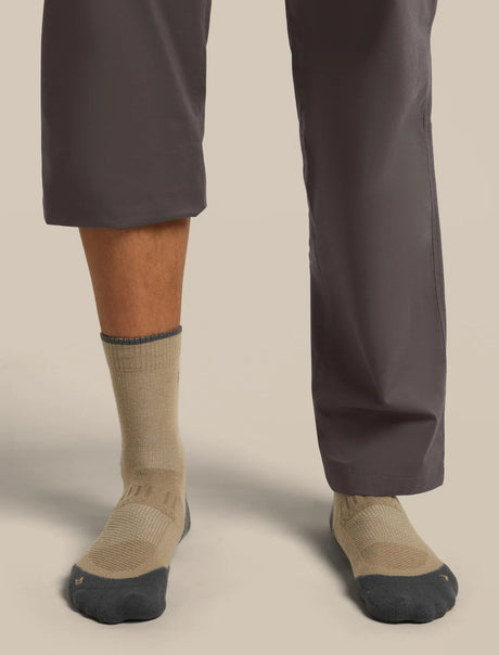 Close-up of a person's lower legs wearing beige crew socks with dark gray toes and heels, paired with dark gray pants, against a solid light beige background. - Icebreaker Merino Hike+ Light Crew Socks (Men's) - Cumin/Obsidian