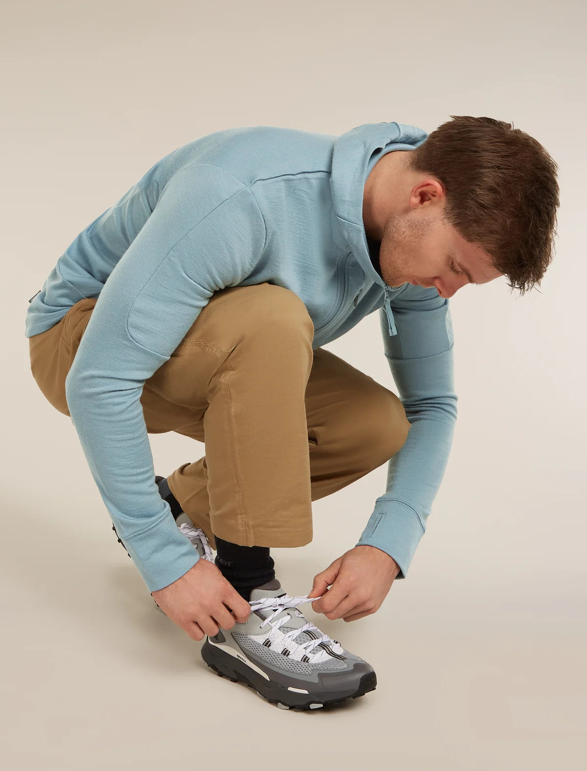 Man in a blue hoodie and beige pants tying his gray and black sneaker on a plain background - Icebreaker Merino Hike+ Heavy Crew Socks (Men's) - Black/Obsidian