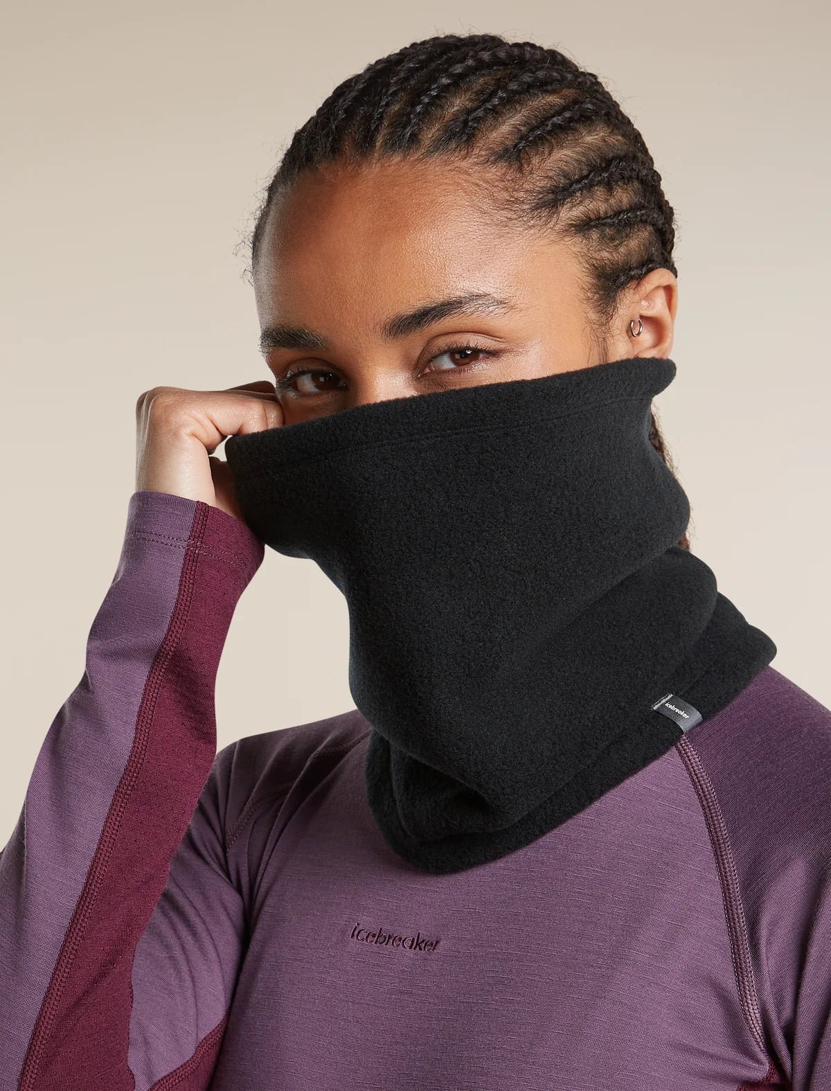 A close-up of a woman wearing a black fleece neck gaiter over her nose and mouth, paired with a purple athletic long-sleeve top. The neck gaiter is pulled up to her chin by her hand. - Icebreaker Merino Blend 800 RealFleece Classic Pile Chute (Unisex) - Black