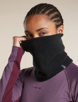 A close-up of a woman wearing a black fleece neck gaiter over her nose and mouth, paired with a purple athletic long-sleeve top. The neck gaiter is pulled up to her chin by her hand. - Icebreaker Merino Blend 800 RealFleece Classic Pile Chute (Unisex) - Black