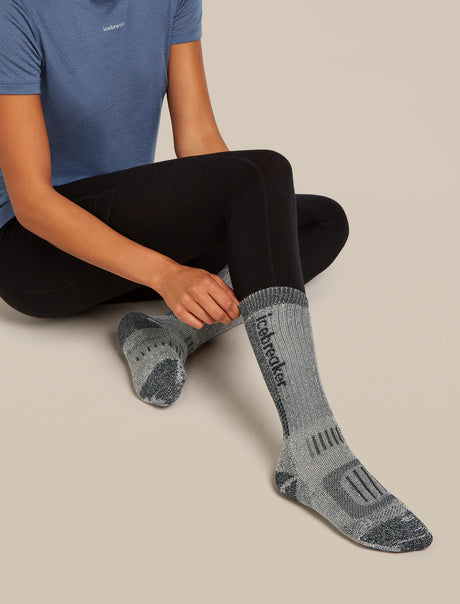 Person wearing gray hiking socks with 'Teva' branding on a beige background - Icebreaker Merino Hike+ Mountaineer Mid Calf Socks (Women's) - Jet Heather/Black