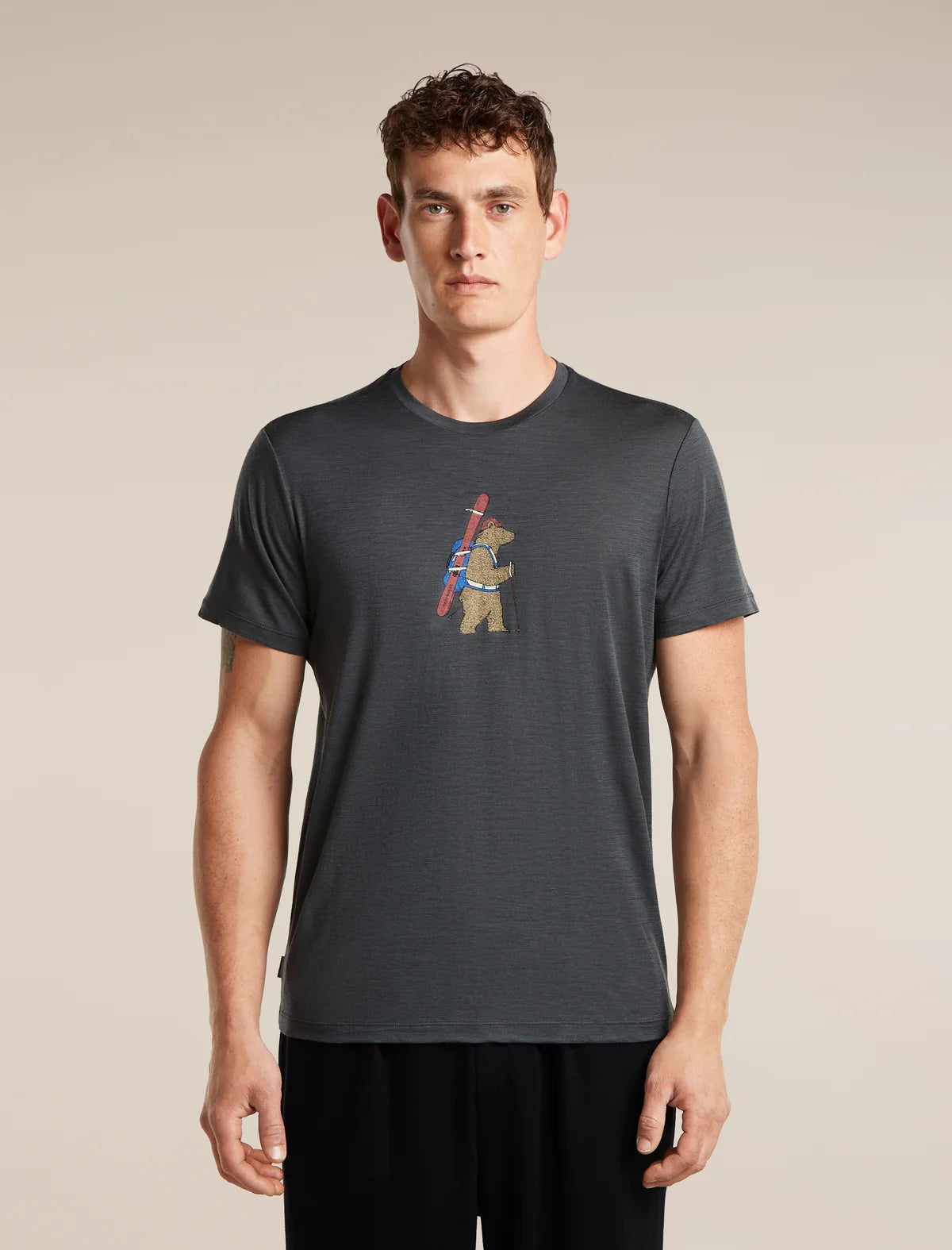 A man wearing a dark gray heathered t-shirt with a small graphic of a bear carrying a ski pole on the chest, standing against a solid beige background. - Icebreaker Merino 150 Tech Lite Short Sleeve T-Shirt Bear Tour (Men's) - Obsidian
