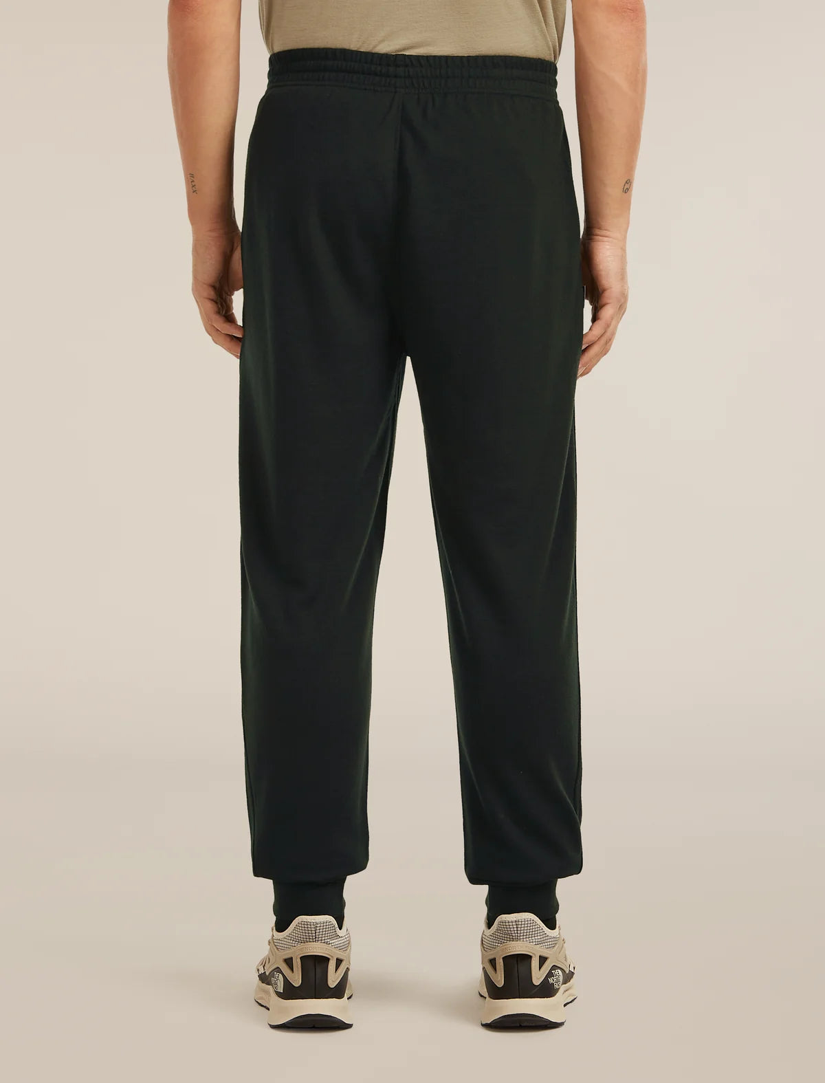 A person shown from the back wearing dark green jogger-style pants with an elastic waistband and cuffs, paired with black socks and beige-and-black sneakers, against a plain light beige studio background. - Icebreaker Merino Blend 150 Drift Pants (Men's) - Black