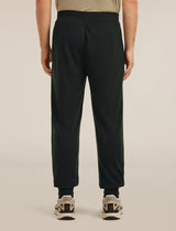 A person shown from the back wearing dark green jogger-style pants with an elastic waistband and cuffs, paired with black socks and beige-and-black sneakers, against a plain light beige studio background. - Icebreaker Merino Blend 150 Drift Pants (Men's) - Black