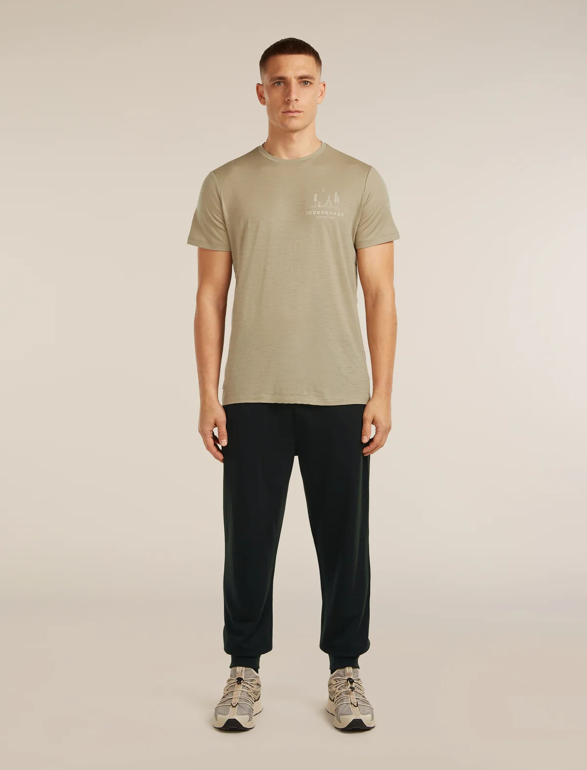 A man modeling a light olive green short-sleeve t-shirt with a small white chest logo, paired with dark navy pants and gray sneakers, against a solid light beige background. - Icebreaker Merino Blend 150 Drift Pants (Men's) - Black