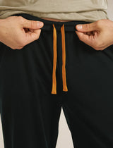 Person adjusting black pants with orange drawstrings on a neutral background - Icebreaker Merino Blend 150 Drift Pants (Men's) - Black