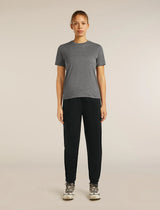 A woman stands against a plain light beige studio background wearing a heather gray short-sleeve crewneck t-shirt, black jogger-style pants, and white and gray sneakers. - Icebreaker Merino Blend 150 Drift Pants (Women's) - Black
