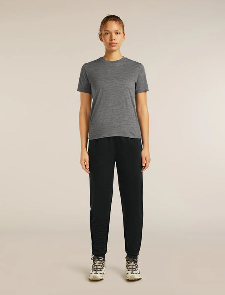 A woman stands against a plain light beige studio background wearing a heather gray short-sleeve crewneck t-shirt, black jogger-style pants, and white and gray sneakers. - Icebreaker Merino Blend 150 Drift Pants (Women's) - Black