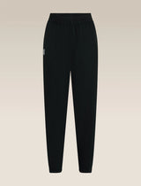 Black jogger-style pants with an elastic waistband and a small white embroidered logo on the left hip, shown against a light beige gradient background. - Icebreaker Merino Blend 150 Drift Pants (Women's) - Black