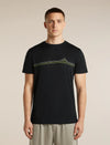 A man wearing a black short-sleeve crewneck t-shirt with a green and yellow mountain graphic across the chest, paired with light gray trousers, set against a plain beige studio background. - Icebreaker Merino 150 Tech Lite Short Sleeve T-Shirt Range Stripes (Men's) - Black