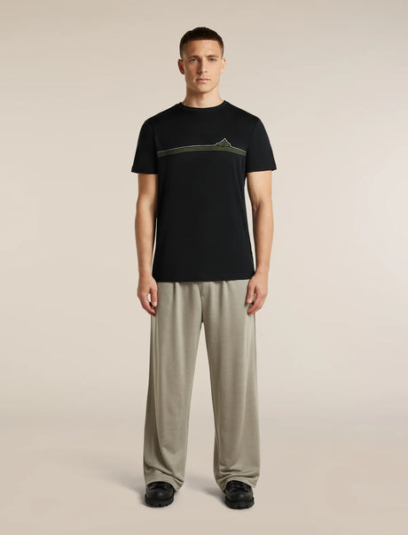 A male model stands against a plain beige studio background wearing a black short-sleeve T-shirt with a horizontal olive-green graphic across the chest and loose light beige pants. - Icebreaker Merino 150 Tech Lite Short Sleeve T-Shirt Range Stripes (Men's) - Black