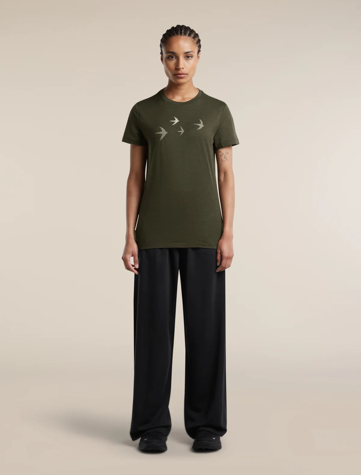 A model stands against a plain beige studio background wearing an olive green short-sleeve t-shirt with a white bird graphic on the chest, paired with black wide-leg trousers. - Icebreaker Merino 150 Tech Lite Short Sleeve T-Shirt Bird Transit (Women's) - Dark Loden