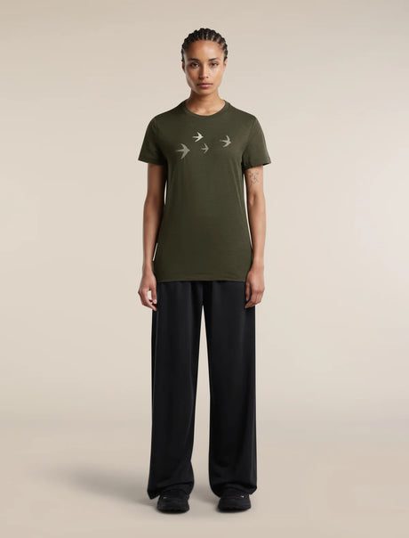 A model stands against a plain beige studio background wearing an olive green short-sleeve t-shirt with a white bird graphic on the chest, paired with black wide-leg trousers. - Icebreaker Merino 150 Tech Lite Short Sleeve T-Shirt Bird Transit (Women's) - Dark Loden