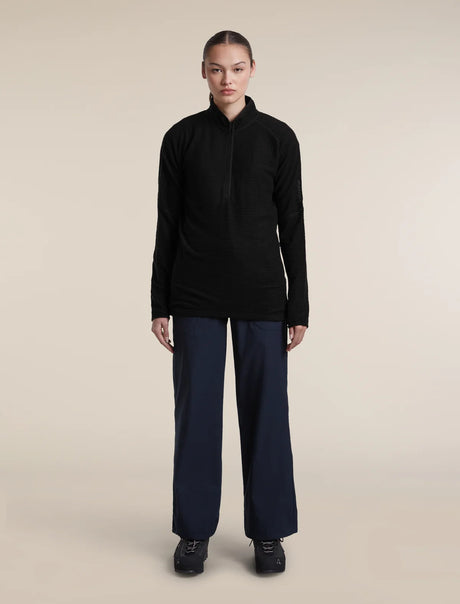 A model stands against a plain beige studio background wearing a black long-sleeve half-zip pullover, dark navy wide-leg trousers, and black shoes. - Icebreaker Merino 400 RealFleece Grid Peaksland Long Sleeve Half Zip (Women's) - Black