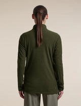 A woman viewed from the back wearing a dark olive green waffle-knit mock-neck sweater and light olive pants, set against a plain beige studio background. - Icebreaker Merino 400 RealFleece Grid Peaksland Long Sleeve Half Zip (Women's) - Dark Loden