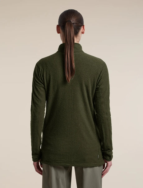 A woman viewed from the back wearing a dark olive green waffle-knit mock-neck sweater and light olive pants, set against a plain beige studio background. - Icebreaker Merino 400 RealFleece Grid Peaksland Long Sleeve Half Zip (Women's) - Dark Loden