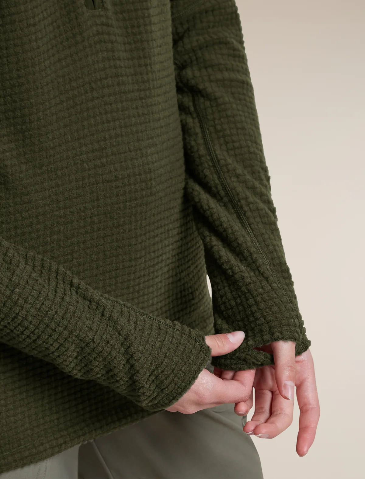 Close-up of a person wearing an olive green waffle-knit sweater, showing the textured fabric and cuffs, with hands adjusting the hem. - Icebreaker Merino 400 RealFleece Grid Peaksland Long Sleeve Half Zip (Women's) - Dark Loden