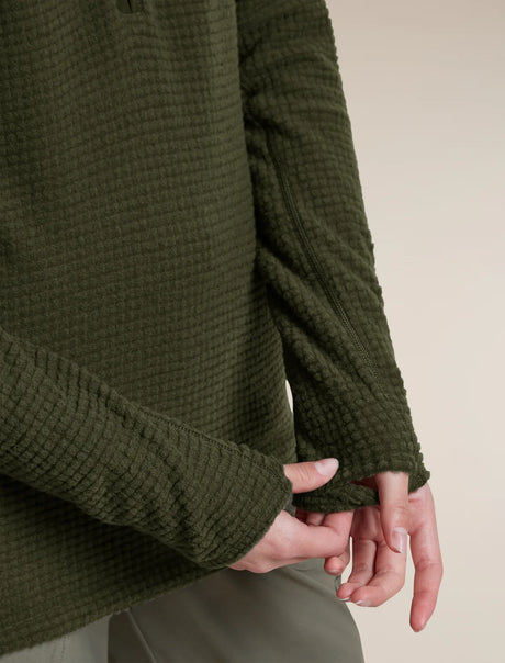 Close-up of a person wearing an olive green waffle-knit sweater, showing the textured fabric and cuffs, with hands adjusting the hem. - Icebreaker Merino 400 RealFleece Grid Peaksland Long Sleeve Half Zip (Women's) - Dark Loden