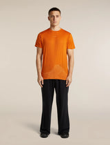 A man modeling a bright orange short-sleeve t-shirt with a subtle tonal mountain graphic near the hem, paired with black trousers, against a solid light beige studio background. - Icebreaker Merino 150 Tech Lite Short Sleeve T-Shirt 3D Mapping (Men's) - Spice