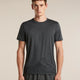 A man wearing a dark gray short-sleeve athletic t-shirt with a subtle mountain line-art graphic near the hem, paired with dark gray athletic pants, against a solid beige background. - Icebreaker Merino 150 Tech Lite Short Sleeve T-Shirt 3D Mapping (Men's) - Obsidian