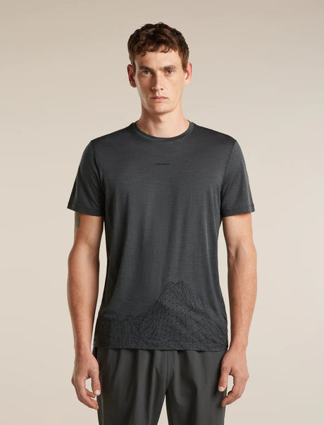 A man wearing a dark gray short-sleeve athletic t-shirt with a subtle mountain line-art graphic near the hem, paired with dark gray athletic pants, against a solid beige background. - Icebreaker Merino 150 Tech Lite Short Sleeve T-Shirt 3D Mapping (Men's) - Obsidian