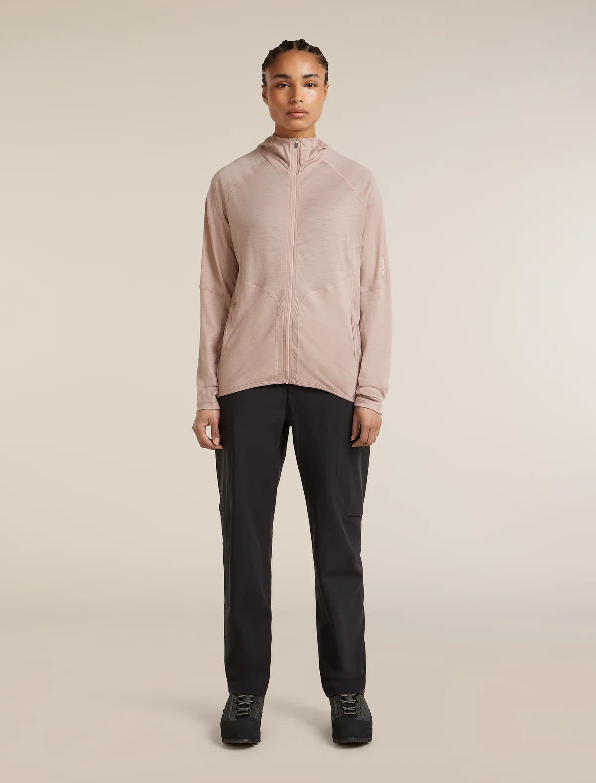 A model stands against a plain beige studio background wearing a light blush pink zip-up athletic jacket, dark charcoal pants, and black sneakers. - Icebreaker 150 MerinoFine Ace Long Sleeve Zip Hoodie (Women's) - Pink Quartz