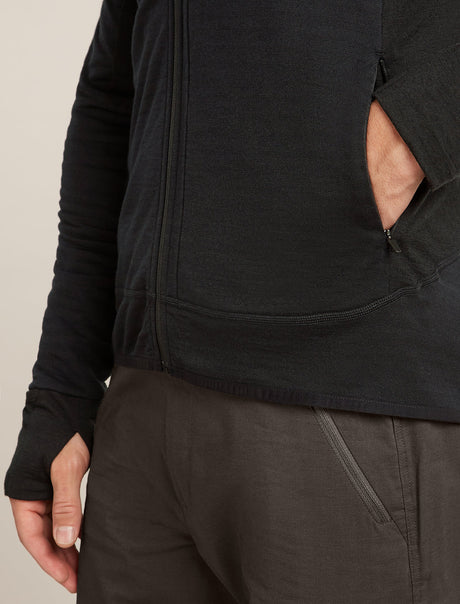 Close-up of a person wearing a black zip-front jacket with a side pocket and dark pants, cropped from the chest down against a plain light beige background. - Icebreaker Merino Blend 300 RealFleece™ Descender Long Sleeve Zip Hoodie (Men's) - Black