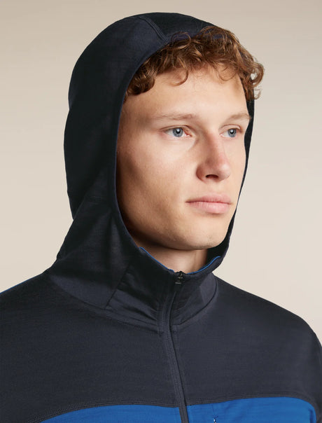 A close-up of a man wearing a dark navy hooded zip-up jacket with a contrasting bright blue panel across the lower chest, set against a plain beige background. - Icebreaker Merino Blend 300 RealFleece™ Descender Long Sleeve Zip Hoodie (Men's) - 
Lazurite/Midnight Navy