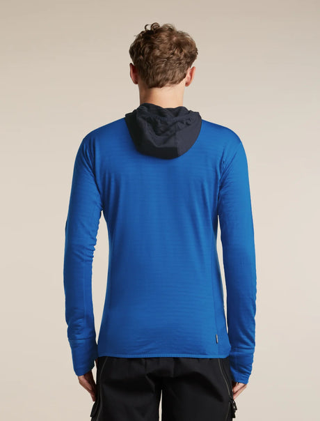 A man viewed from behind wearing a bright blue long-sleeve athletic hoodie with a dark navy hood, paired with black pants, against a solid beige background. - Icebreaker Merino Blend 300 RealFleece™ Descender Long Sleeve Zip Hoodie (Men's) - 
Lazurite/Midnight Navy