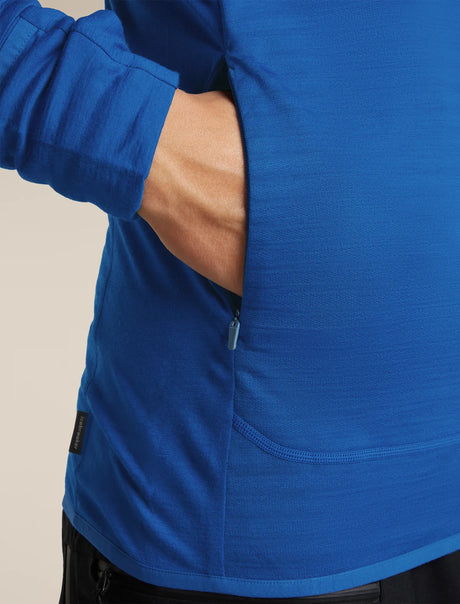 Close-up of a person wearing a bright blue long-sleeve athletic top, showing a side pocket with a zipper and the arm tucked into the pocket, against a plain light beige background. - Icebreaker Merino Blend 300 RealFleece™ Descender Long Sleeve Zip Hoodie (Men's) - 
Lazurite/Midnight Navy