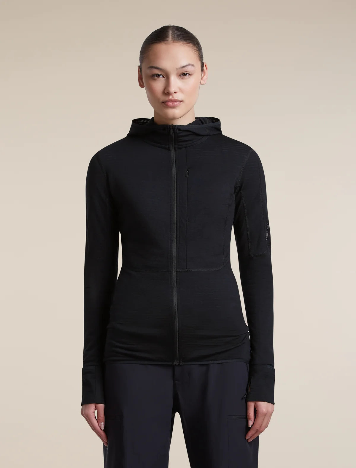 A woman modeling a black long-sleeve athletic zip-up hoodie with a hood, shown against a plain beige studio background. - Icebreaker Merino Blend 200 RealFleece Descender Long Sleeve Zip Hoodie (Women's) - Black
