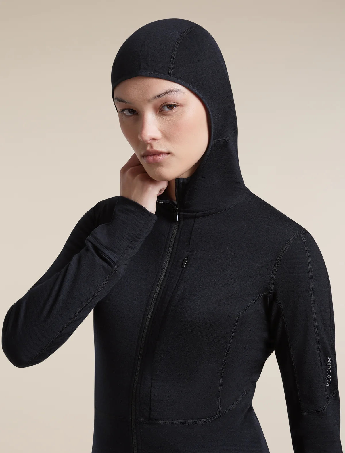 A woman wearing a black hooded athletic jacket with a full front zipper, photographed against a plain warm beige studio background. - Icebreaker Merino Blend 200 RealFleece Descender Long Sleeve Zip Hoodie (Women's) - Black